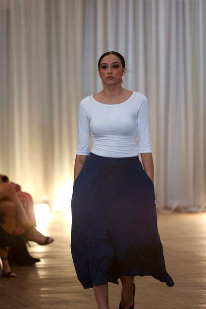 This is a young woman facing the camera silhouetted against a cream drape background. Her hair is tied back, she wears a cream 3/4 length sleeve top with a scoop neck. She wears a our blue Boho Skirt with is which has pockets and a deep gathered frill.