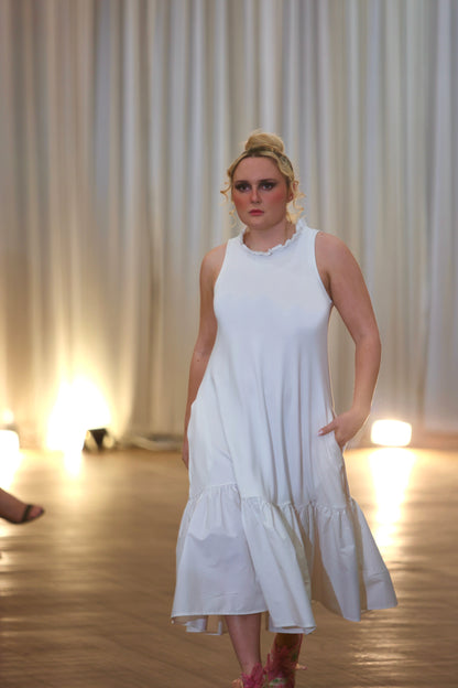 There is a young woman walking towards the camera in a well lit room with long white curtains in the background. She has long blonde hair tied up. She is wearing the iris midi dress with pockets in cream. The dress has a high neck with a row of ruffles. She is wearing red shoes and her left hand is in her pocket. Her right hand is by her side.