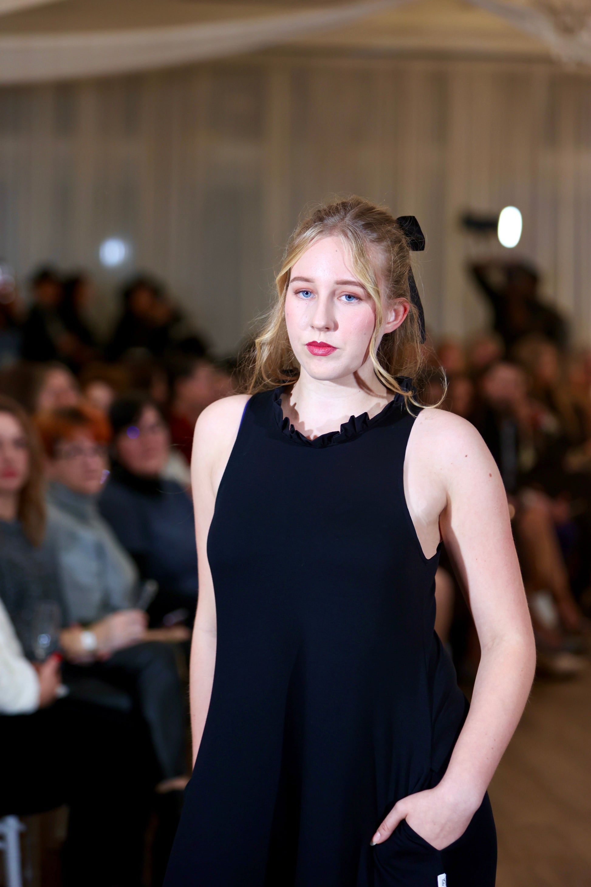 There is a young woman walking towards the camera in a crowded room. She has long blonde hair tied in a bow. She is wearing the iris midi dress with pockets in black. The dress has a high neck with a row of ruffles. Both hands are in her pockets.