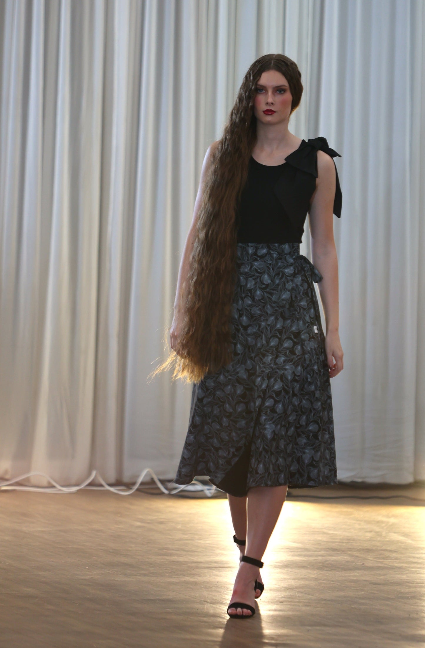 The young woman in the picture has very long dark hair and she is walking towards the camera in a well lit room. She is wearing a brook top with bow in black. She is also wearing our boho gathered midi skirt with pockets in navy print. She is wearing black shoes. Her right hand is hidden by her long hair while her left hand is by her side.