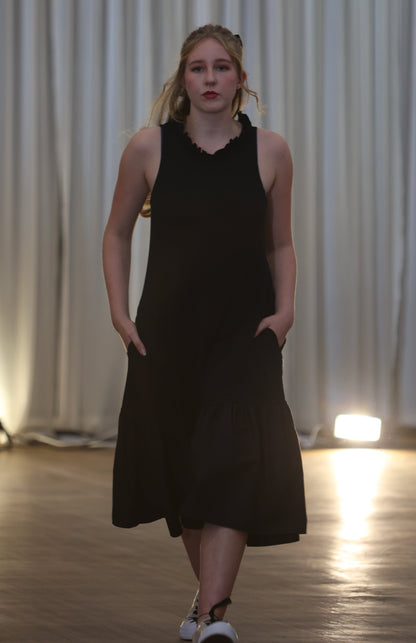 There is a young woman walking towards the camera in a well lit room with long white curtains in the background. She has long blonde hair. She is wearing the iris midi dress with pockets in black.  The dress has a high neck with a row of ruffles. She is wearing white shoes and both hands are in her pockets.